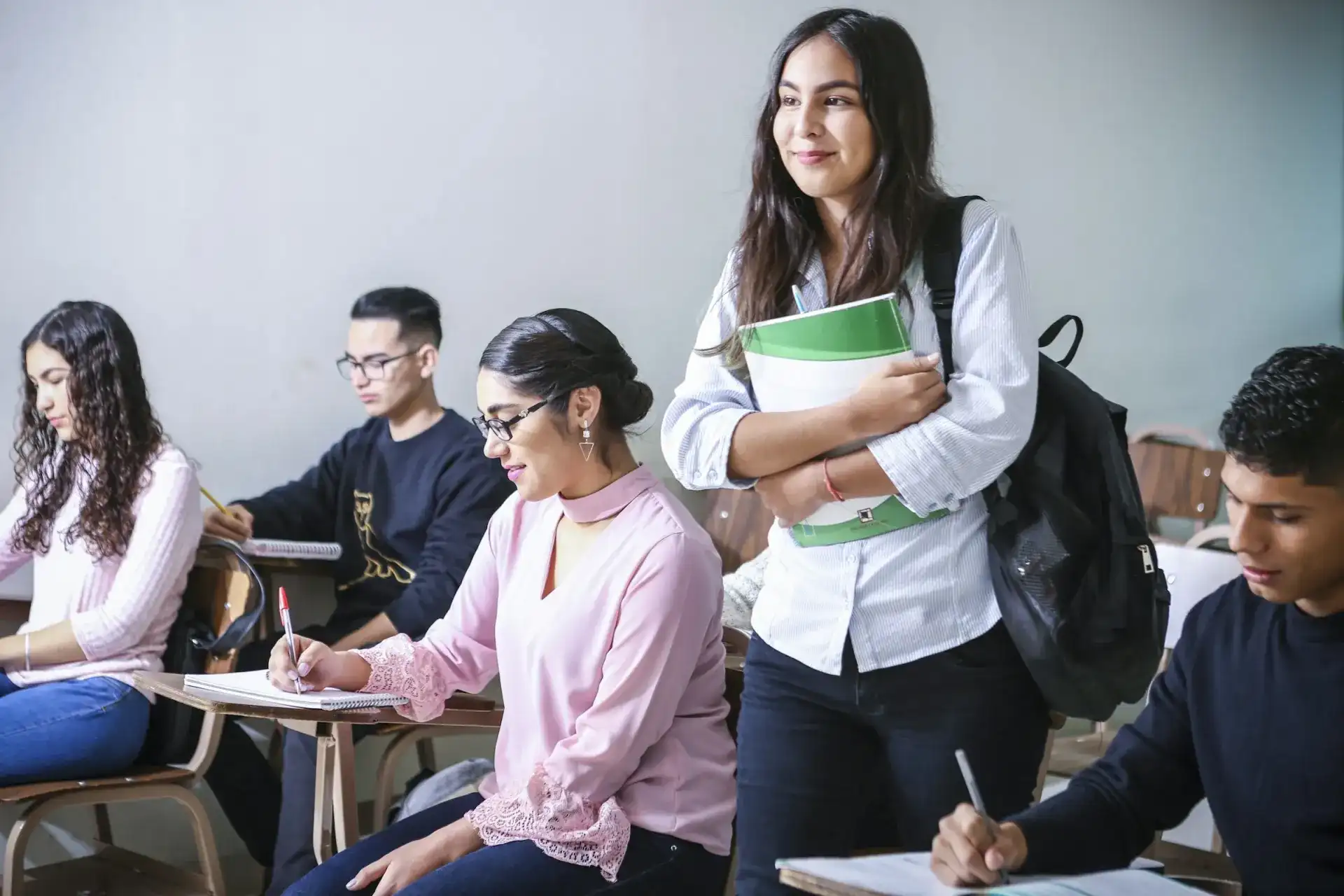 Students seated at wooden desks in a classroom, one young woman standing with a green textbook and a backpack