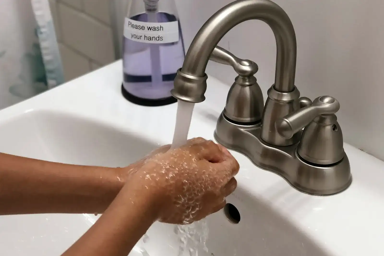 Hands washing with soap under a running faucet beside a bottle labelled Please wash your hands
