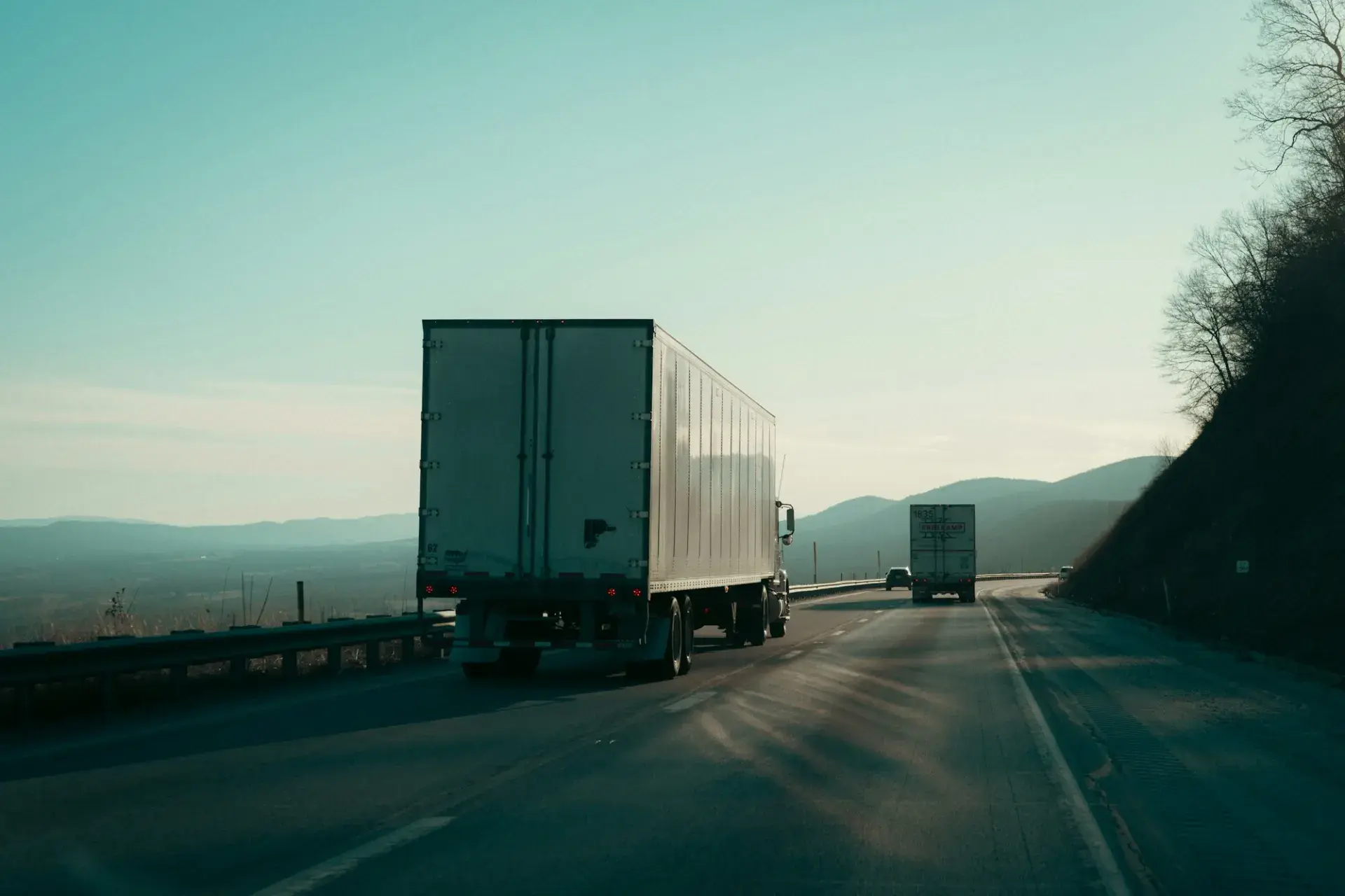 A box-trailer truck climbing a mountain highway at dusk with another truck ahead