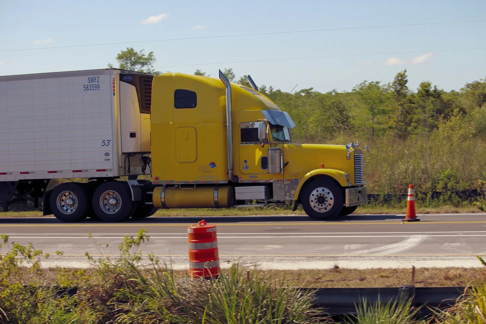 A yellow tractor-trailer driving along a highway past orange traffic barrels
