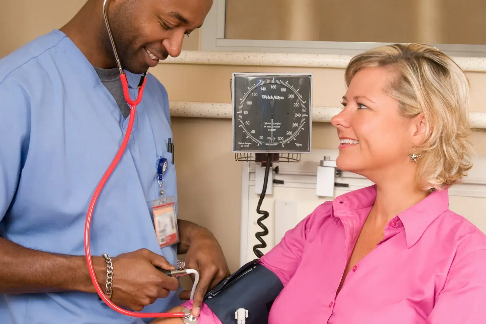 Healthcare worker in blue scrubs taking a smiling patient's blood pressure with a wall gauge and stethoscope