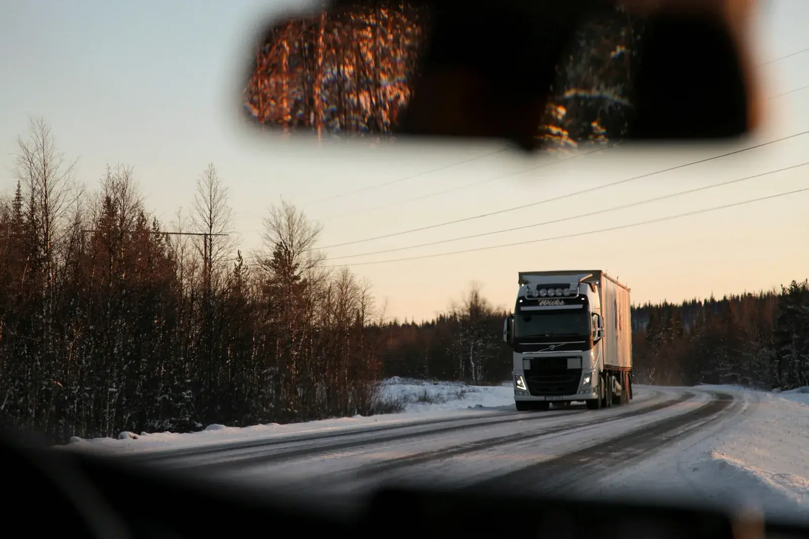 Semi truck coming around a snowy curve, seen through a car windshield at dusk