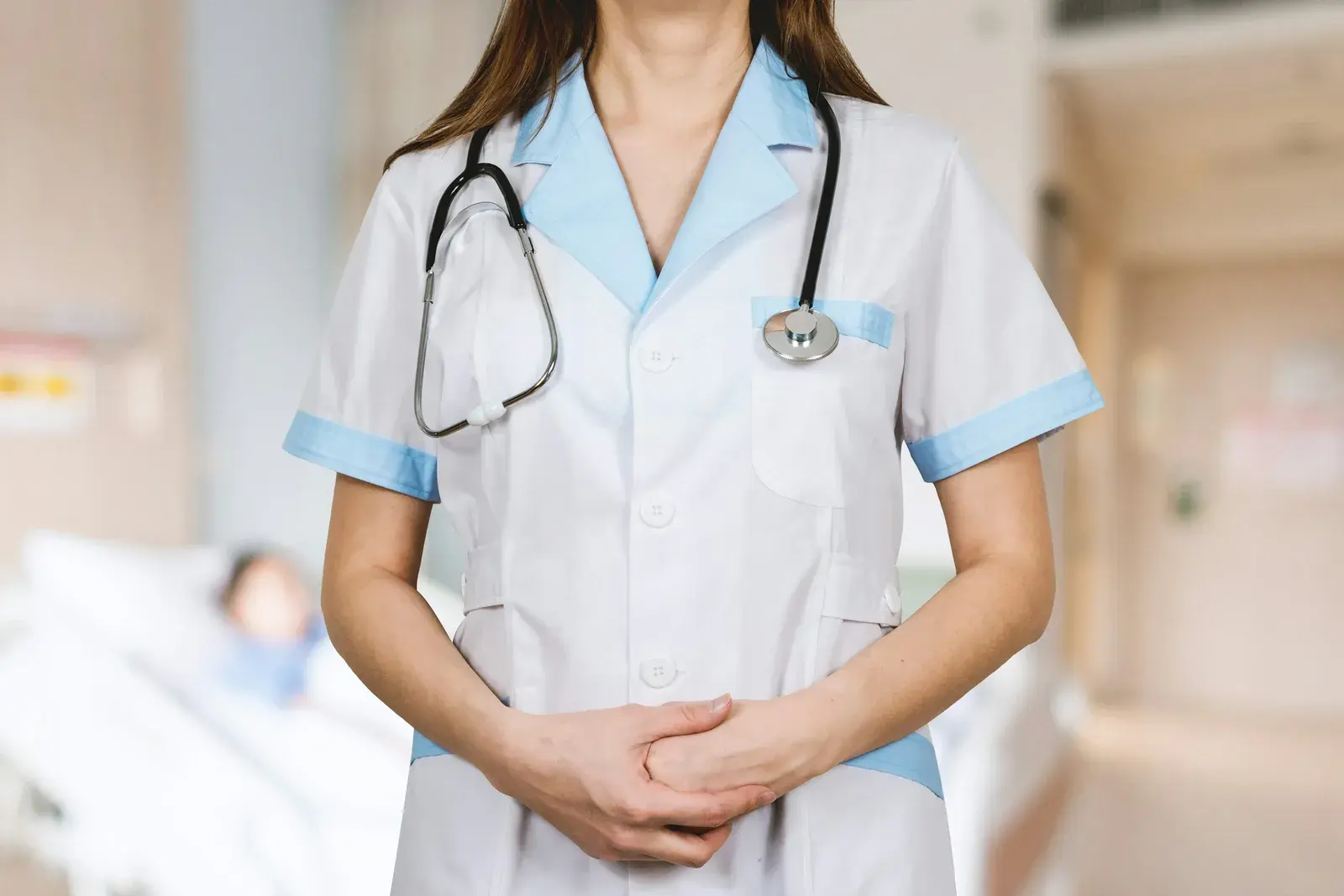 Nurse in a white uniform with a stethoscope standing in a hospital room, a patient in bed behind her